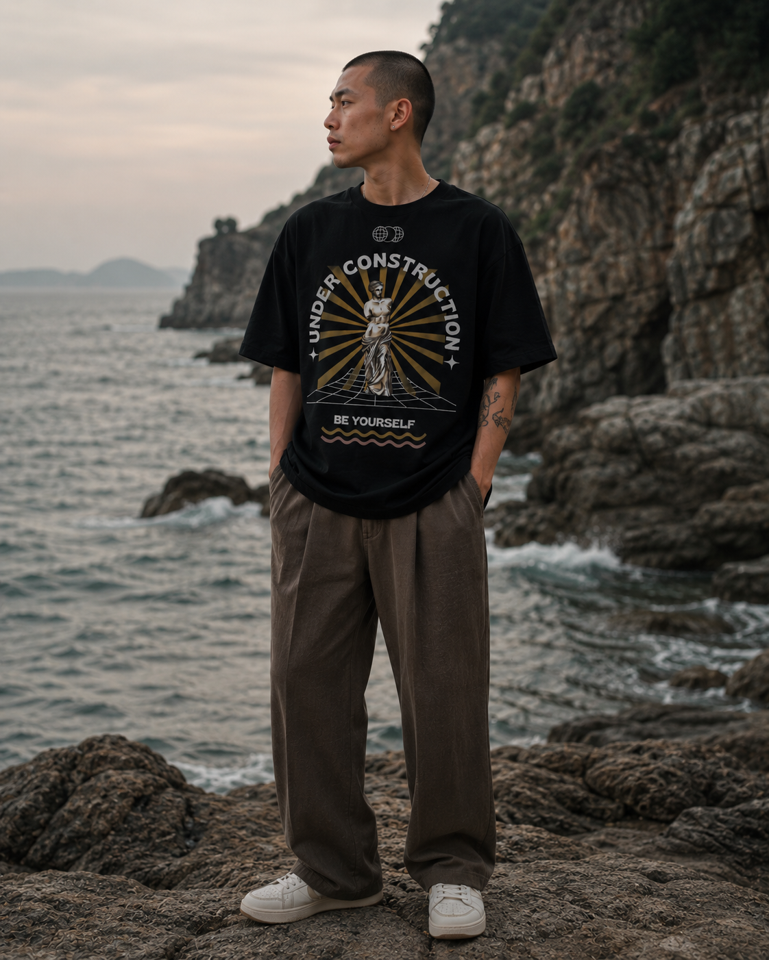 Man wearing a black t-shirt with graphic design standing on rocky terrain by the sea. Stylist streetwear tee, comfy formal tee, fashion tee, graphic tee