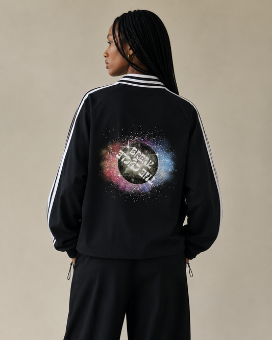 Person wearing a black tracksuit with a cosmic design on the back against a beige background. Keep the same model and background as the previous image, but show the back of the sweatshirt