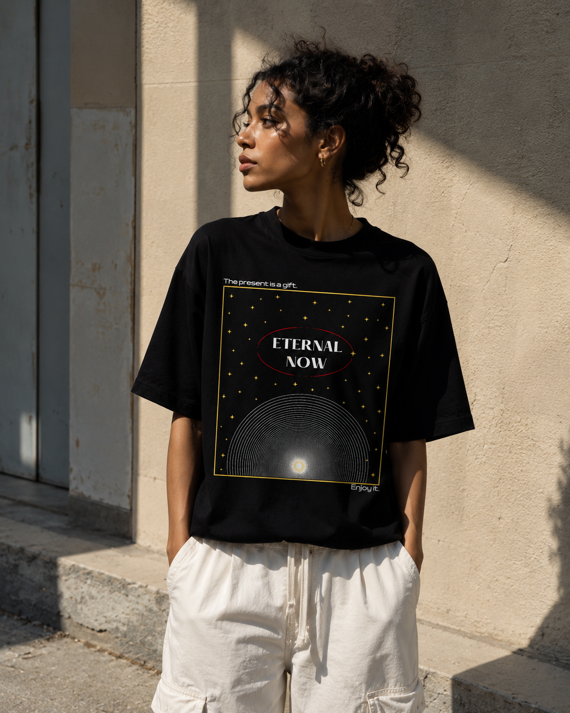 Person wearing a black t-shirt with 'Eternal Now' design against a beige wall. Stylist streetwear tee, comfy formal tee, fashion tee, graphic tee