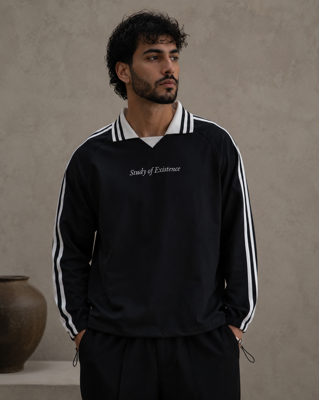 Man wearing a black tracksuit with white stripes and 'Study of Existence' text, standing against a beige wall. Stylist streetwear collar Sweatshirt, comfy formal collar Sweatshirt, fashion collar Sweatshirt, graphic collar Sweatshirt