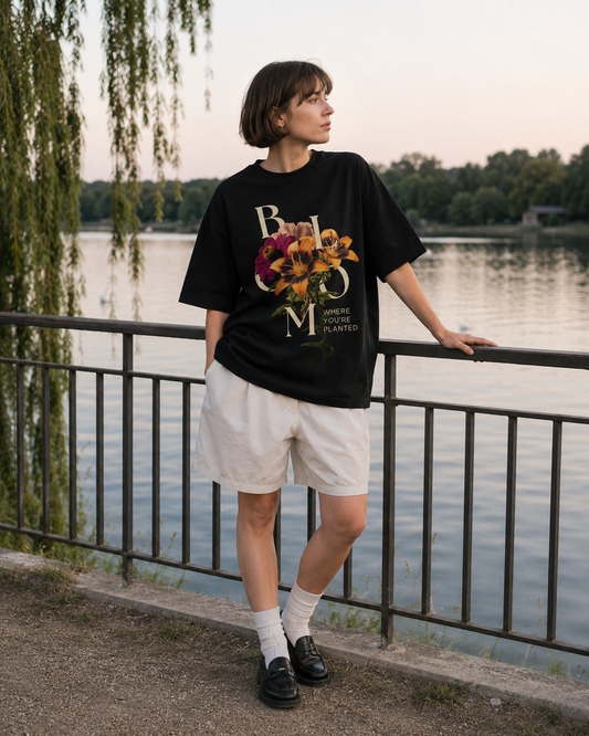 Person wearing a black t-shirt with floral design and text, standing by a lake. Stylist streetwear tee, comfy formal tee, fashion tee, graphic tee