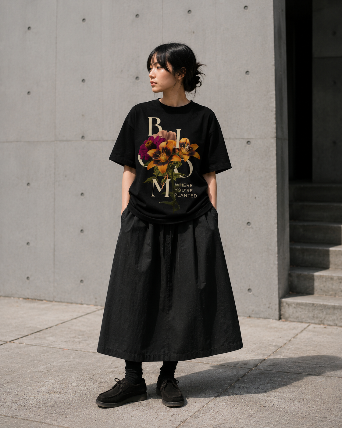 Person wearing a black t-shirt with colorful floral design and text, paired with a long black skirt, standing against a concrete wall. Stylist streetwear tee, comfy formal tee, fashion tee, graphic tee