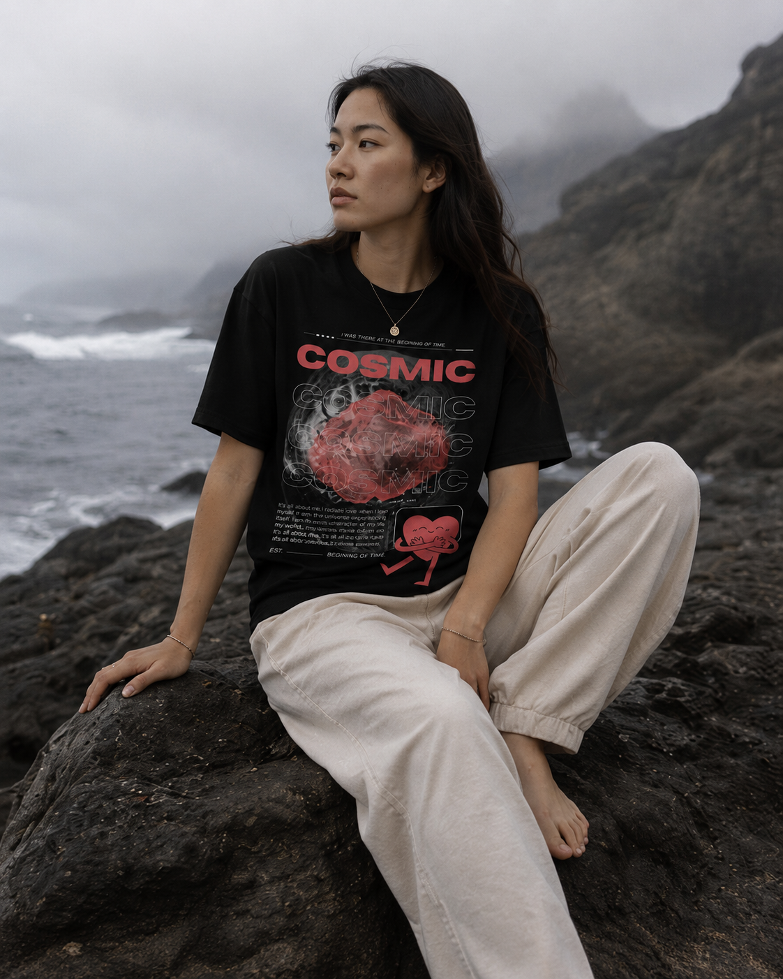 Person wearing a black t-shirt with 'Cosmic' design sitting on rocks by a body of water. Stylist streetwear tee, comfy formal tee, fashion tee, graphic tee