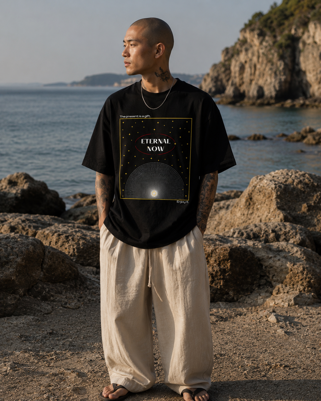 Person wearing a black t-shirt with 'Eternal Now' design by the ocean. Stylist streetwear tee, comfy formal tee, fashion tee, graphic tee
