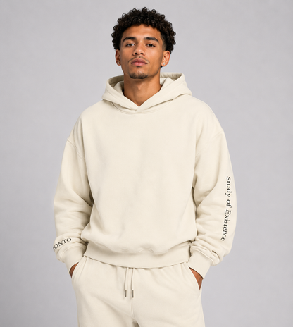 Man wearing a beige hoodie and pants with text on sleeves against a gray background. Stylist streetwear hoodie, comfy formal hoodie, fashion hoodie, graphic hoodie, no front pocket hoodie, large hooded hoodie, large hood hoodie, oversized hoodie