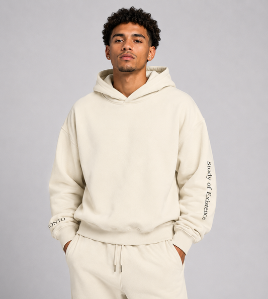 Man wearing a beige hoodie and pants with text on sleeves against a gray background. Stylist streetwear hoodie, comfy formal hoodie, fashion hoodie, graphic hoodie, no front pocket hoodie, large hooded hoodie, large hood hoodie, oversized hoodie