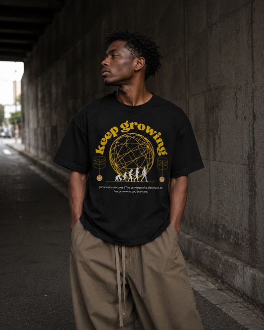 Man wearing a black t-shirt with 'keep growing' text and graphic, standing against a concrete wall. Stylist streetwear tee, comfy formal tee, fashion tee, graphic tee