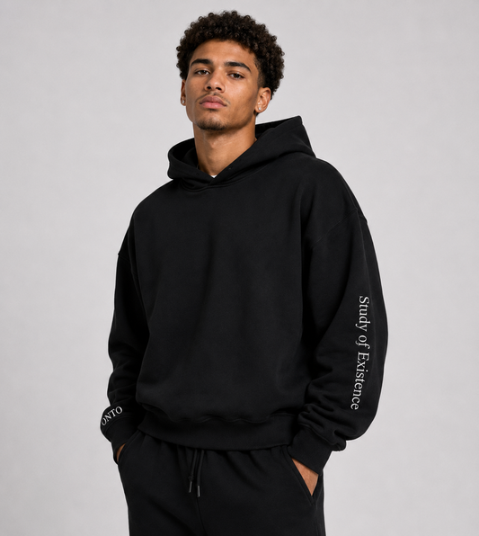 Man wearing a black hoodie with text on sleeves against a gray background. Stylist streetwear hoodie, comfy formal hoodie, fashion hoodie, graphic hoodie, no front pocket hoodie, large hooded hoodie, large hood hoodie, oversized hoodie