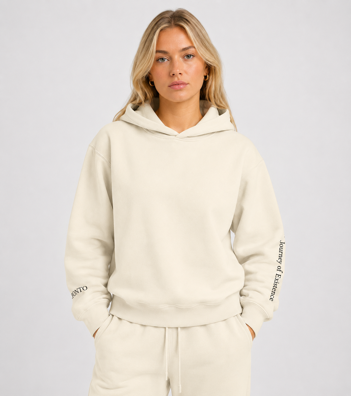 Person wearing a beige hoodie with branding on sleeves against a plain background. Stylist streetwear hoodie, comfy formal hoodie, fashion hoodie, graphic hoodie, no front pocket hoodie, large hooded hoodie, large hood hoodie, oversized hoodie